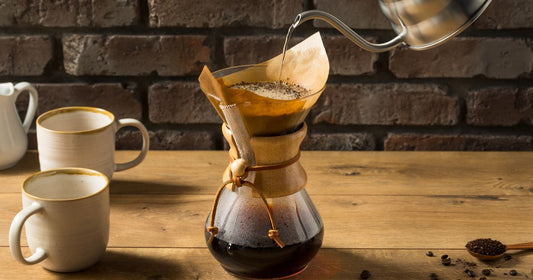 Pour-over coffee brewing into a glass carafe on a wooden table with coffee mugs and roasted coffee grounds nearby